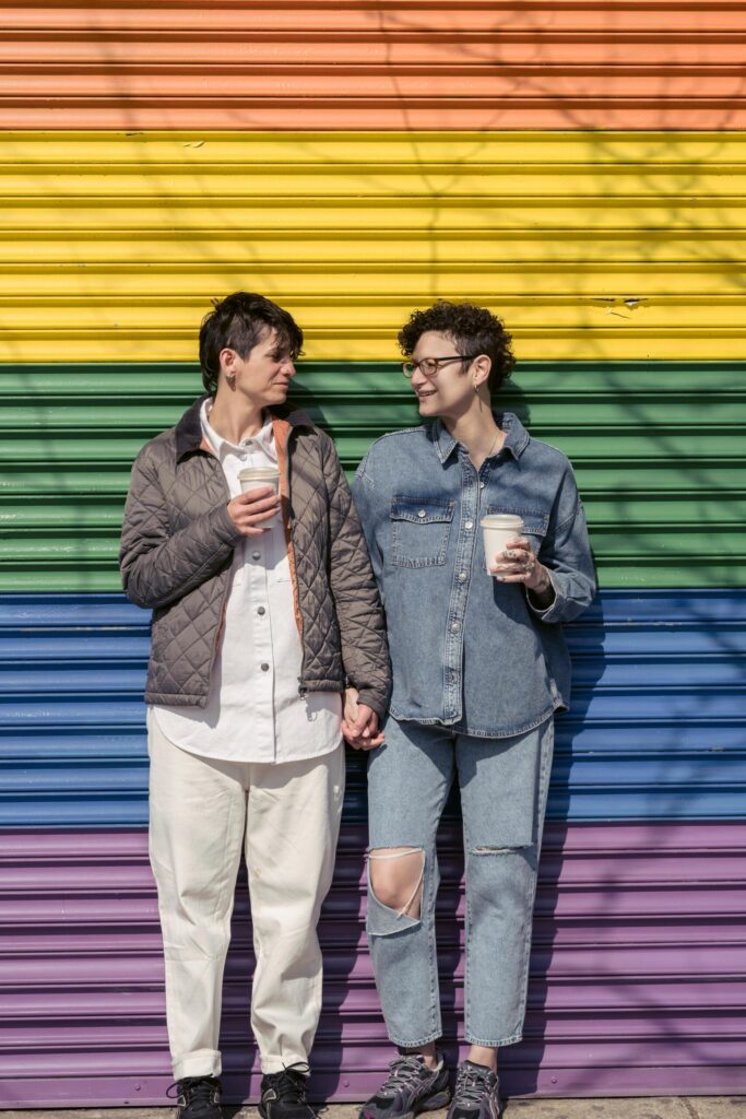 Full body of content lesbians with takeaway drinks looking at each other while standing on colorful background with LGBT rainbow flag on sunny street